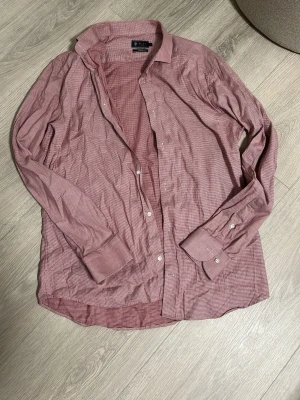 Rutig röd skjorta från Riley - Säljer en röd och vit rutig skjorta från Riley. Skjortan har klassisk krage, långa ärmar med manschetter och vita knappar framtill. Tillverkad i mjuk bomull och har en avslappnad passform. Perfekt för dig som gillar stilrena detaljer.