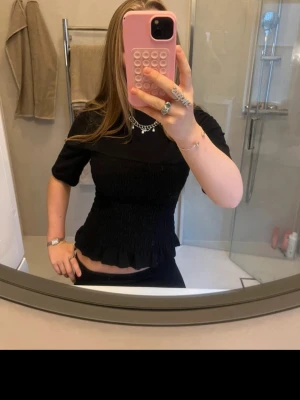 Svart croppad topp med volangkant - Säljer en svart croppad topp med korta ärmar och volangkant nedtill. Toppen har en smockad, åtsittande passform som ger en snygg siluett. Perfekt att styla med jeans eller kjol för en trendig look.