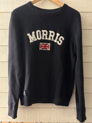 Mörkblå stickad tröja Morris - Mörkblå stickad tröja från Morris med broderad logga och brittisk flagga på bröstet. Tröjan har rund halsringning med ribbade detaljer samt långa ärmar och ribbade muddar. Perfekt för en stilren och klassisk look.