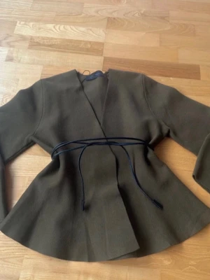 Olivgrön omlottkavaj från Zara - Snygg olivgrön stickad kofta från Zara med knytband i midjan. Koftan har lång ärm och en lätt utsvängd form nertill. Perfekt för dig som gillar stilrena och trendiga plagg.