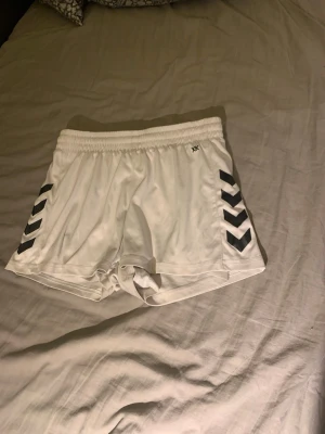 Vita sportshorts från Hummel - Säljer ett par vita sport soprtshorts från Hummel med svarta chevron-detaljer på sidorna och elastisk midja. Perfekta för träning eller sportaktiviteter. Klassisk Hummel-logga på ena benet och mjukt, lätt material.