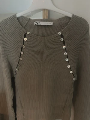 Beige  topp  från Zara - Snygg beige  stickad tröja från Zara med dekorativa silverfärgade knappar längs framsidan. Tröjan har rund halsringning och är ribbstickad för en tightare passform därav sitter den som S. Perfekt för dig som gillar unika detaljer och trendig stil.