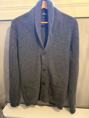 Mörkgrå stickad kofta med sjalkrage - Knappt använd mörkgrå stickad kofta från Dressmann med sjalkrage och knäppning framtill. Två stora fickor framtill och ribbade muddar vid ärmslut och nederkant. Perfekt för lager på lager och chill stil. Knappt använd. Nypris 700