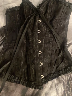 Svart korsett med spetsdetaljer - Snygg svart korsett med glansiga broderade mönster och volangkant upptill och nedtill. Framsidan har metallknäppning och baksidan snörning för perfekt passform. Materialet är glansigt tyg med dekorativa detaljer och satinband. Köpt secondhand, så vet tyvärr inte vart den är ifrån. Har ett litet hål, är inte synligt när den är på.