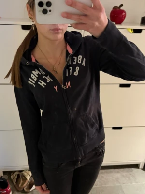Mörkblå Abercrombie & Fitch zip up hoodie  - Zip up hoodie med dragkedja från Abercrombie & Fitch. Har vit och rosa text på bröstet. Perfekt för en avslappnad och cool stil. Storlek L men passar S bättre 