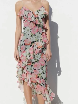 Blommig volangklänning med smala axelband - Superfin klänning med blommigt mönster i rosa och grönt på ljus botten. Klänningen har smala axelband, v-ringning och flera volangdetaljer längs kjolen och överdelen. Perfekt för sommarens alla tillfällen.