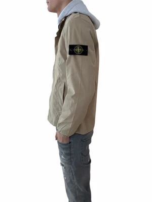 Beige vindjacka från Stone Island - Storlek XL passar L. Säljer en beige vindjacka från Stone Island med klassisk patch på vänster ärm. Jackan har dragkedja framtill, elastiska muddar och en stilren design. Perfekt för vår och höst, lätt att matcha med olika outfits. Passformen är normal och jackan är långärmad.