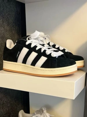 Adidas Campus svartvita sneakers - Klassiska Adidas Campus sneakers i svart mocka med vita detaljer och de ikoniska tre ränderna på sidan. Skorna har vit sula, rund tå och snörning. Perfekta för en stilren och sportig look.