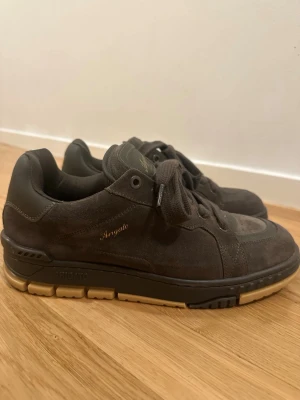 Bruna sneakers från Axel Arigato - Snygga bruna sneakers från Axel Arigato i mocka med gulddetaljer och tjock sula. Skorna har snörning och en låg profil, perfekt för en stilren och modern look. Axel Arigato-logga syns på sidan och plösen.