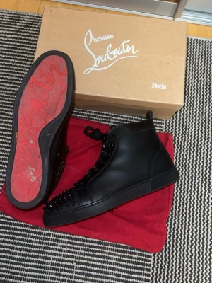 Svarta Christian Louboutin sneakers - Snygga svarta high-top sneakers från Christian Louboutin med ikoniska röda sulor och svarta nitar på tån. Skorna är i skinn och har snörning framtill. Perfekta för dig som vill sticka ut med en lyxig och edgy stil. Tillkommer Box och Dustbags, vid frågor är det bara att höra av sig