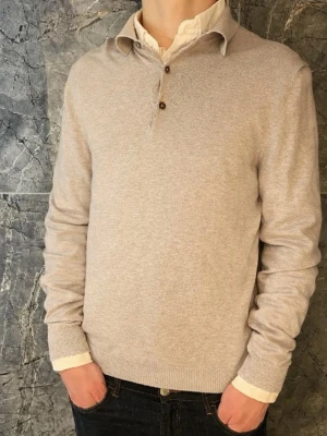 Beige stickad tröja från Bläck - Snygg beige stickad tröja från Bläck med krage och knappar vid halsen. Tröjan har långa ärmar och ribbade muddar vid ärmslut och nederkant. Perfekt för lager-på-lager och passar till både skjorta och t-shirt.