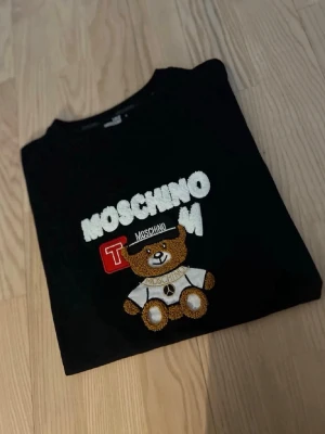 Svart Moschino T-shirt med nalle - Cool svart T-shirt från Love Moschino med broderad nalle och logga framtill. T-shirten har rund hals och korta ärmar, perfekt för en trendig och lekfull stil. Materialet är mjukt och bekvämt, passar perfekt till jeans eller kjol.