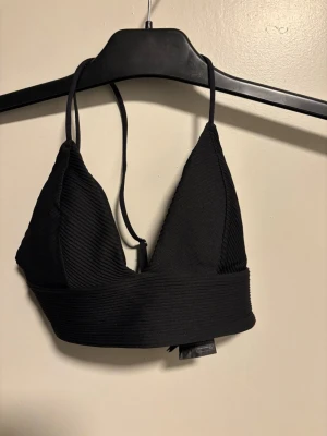 Svart ribbad bikini överdel med smala band - Svart bikiniöverdel med ribbad struktur och triangelformade kupor. Smala axelband som korsas i ryggen och bred resår under bysten för extra stöd. Perfekt för en stilren och bekväm look.