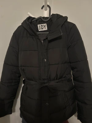 Svart pufferjacka från JDY med huva - Svart pufferjacka från JDY med huva och knytbälte i midjan. Jackan har quiltad design, dragkedja och tryckknappar framtill. Perfekt för kyliga dagar och har en klassisk, enkel look.