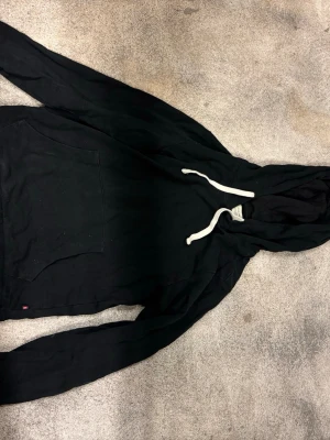 Svart hoodie från Jack & Jones XS - Svart hoodie från Jack & Jones med klassisk känguruficka och vita dragsnören i huvan. Tillverkad i mjuk bomullsmix, perfekt för en avslappnad stil. Långärmad och enkel design som funkar till det mesta.