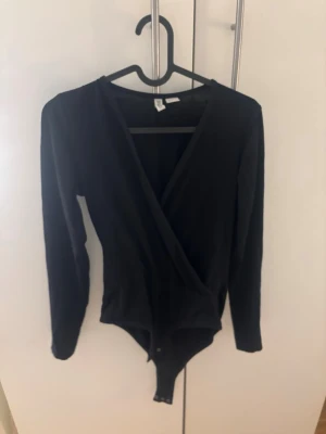 Svart omlottbody med långa ärmar - Snygg svart body med omlott-skärning och långa ärmar. Bodyn har tryckknappar i grenen och är tillverkad i ett mjukt, stretchigt material som sitter skönt på kroppen. Perfekt att styla med jeans eller kjol för en trendig look. Bodyn är i storlek S men är för stor för mig så skulle säga mer M