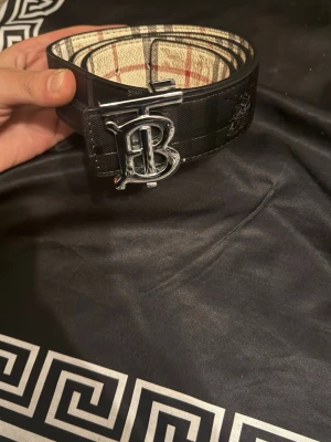 Burberry svart bälte  - Snyggt svart bälte från Burberry med klassiskt rutigt innerfoder och ett stort silverfärgat B'. Bältet har en stilren design med diskret mönster på utsidan och är tillverkat i läder.