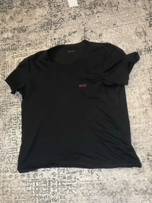  t-shirt  BOSS - Svart t-shirt från BOSS med diskret röd logga på bröstet. Klassisk rund halsringning och korta ärmar. Perfekt basplagg med stilren design, tillverkad i mjuk bomull för en bekväm känsla. Storlek L men passar perfekt för mig som är M