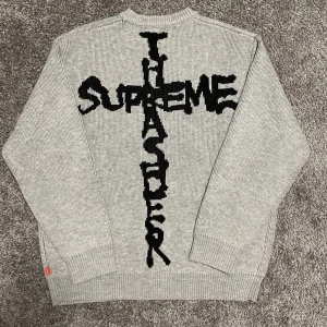 Grå Supreme stickad tröja  - Stickad tröja i ljusgrått från Supreme. Tröjan har ett stort svart korsformat Supreme. Rund halsringning och ribbade muddar. Perfekt för kalla dagar. Använd en gång och är i storlek S