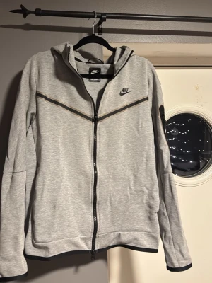 Grå Nike tech hoodie med dragkedja - Snygg grå hoodie från Nike med hel dragkedja framtill och svart Nike-logga på bröstet. Storlek M. Använd ett fåtal gånger