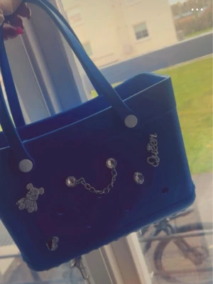 Blå handväska med kedjedetaljer - Cool blå handväska i syntetmaterial med silverfärgade detaljer som kedja, nitar och en liten björn. Väskan har två handtag och en stilren, boxig form. Perfekt för dig som vill sticka ut med accessoarer som syns.