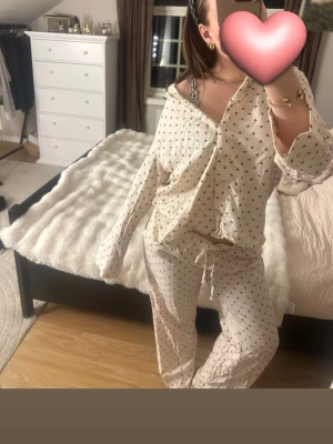 Vit pyjamas med röda hjärtan - Säljer en söt tvådelad pyjamas i beige med små röda hjärtan över hela. Skjortan har knappar framtill och långa ärmar, byxorna har resår och dragsko i midjan. Perfekt för mysiga kvällar hemma.