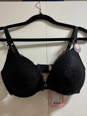 Svart spets-bh från Hunkemöller - Snygg svart bh från Hunkemöller med vadderade kupor och vacker spetsdetalj över hela framsidan. Justerbara axelband och klassisk hyskknäppning bak. Liten rosett och dekorativ ring framtill ger extra detalj. Perfekt för dig som gillar stilrena underkläder.
