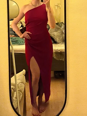 Vinröd oneshoulder långklänning - Säljer en elegant vinröd långklänning med oneshoulder-design och hög slits på ena sidan. Klänningen har en figurnära passform och är perfekt för dig som vill sticka ut. Materialet känns mjukt och följsamt mot kroppen.