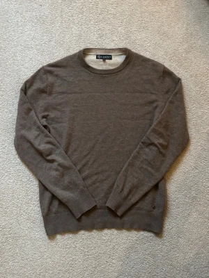 Stickad tröja Lagency - Klassisk brun stickad tröja från Oscar Jacobson i tunn ullmix. Tröjan har rund halsringning, långa ärmar och ribbade muddar vid ärmslut och nederkant. Perfekt för lager på lager och enkel att matcha med olika stilar.