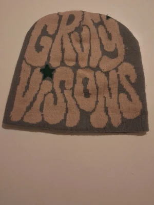 Grå mössa med tryck och spindelnät - Säljer en grå stickad Gravity Vision mössa med beige text och en grön stjärna på framsidan. Baksidan har ett coolt spindelnätsmönster i beige. Perfekt accessoar för dig som vill sticka ut.