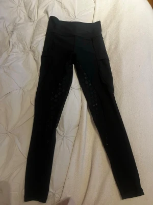 Mayadelorez ridbyxor - Snygga svarta ridleggings med hög midja och praktiska fickor på sidorna. Silikonmönster på insidan av benen för extra grepp i sadeln. Perfekta för dig som vill ha både stil och funktion i stallet.