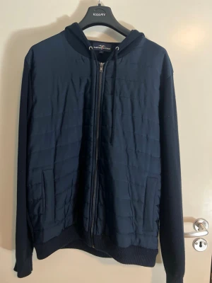 Mörkblå stickad cardigan med luva och dragkedja - Snygg mörkblå hoodie från Hampton republic med dragkedja framtill och två fickor. Utsidan har quiltad struktur och luvan har dragsko. Ärmar och baksida är i mjukare tyg, medan framsidan är vadderad för extra värme. Perfekt för en chill och stilren look.