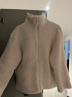 Beige teddyjacka med dragkedja - Mysig beige teddyjacka med hög krage och hel dragkedja framtill. Jackan har en loose passform och är tillverkad i fluffigt teddy-material som håller dig varm och bekväm. Perfekt för kyliga dagar och enkel att matcha med olika outfits.