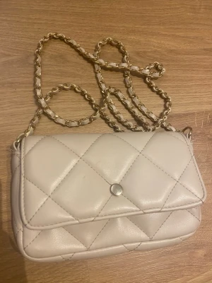 Beige quiltad handväska med kedja - Säljer en beige quiltad handväska med guldfärgad kedjerem och magnetlås. Väskan har en klassisk rektangulär form och är tillverkad i syntetmaterial. Perfekt för dig som gillar stilrena detaljer och trendig design.