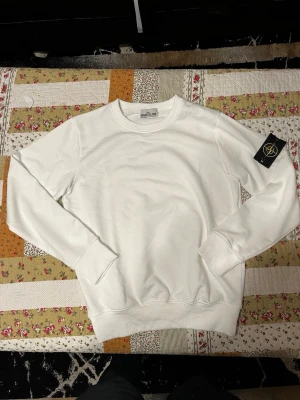 Stone island tröja - Säljer en clean vit sweatshirt från Stone Island med den klassiska svarta patchen med gul kompass på vänster ärm. Tröjan har rund halsringning, ribbade muddar och är tillverkad i mjukt bomullsmaterial. Perfekt för en stilren och avslappnad look. Helt ny 