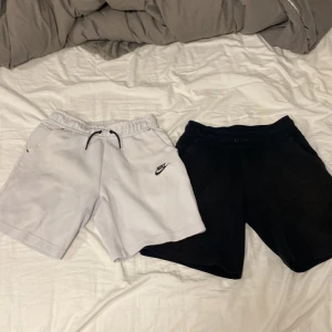 Nike tech shorts i svart och vitt - Två par Nike tech shorts, ett par vita med svart snörning och logga, samt ett par svarta. Båda har elastisk midja och sidofickor, perfekta för en avslappnad stil eller träning. De är i bra skick och perfekta för sommaren som kommer om några månader!