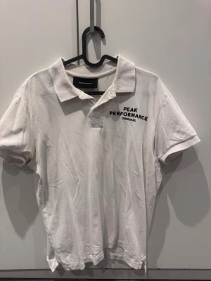 Vit pikétröja Peak Performance - Klassisk vit pikétröja från Peak Performance med svart logga broderad på bröstet. Tröjan har korta ärmar, krage och knappar framtill. Mycket bra skick!!