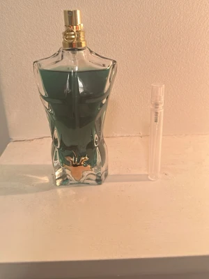 Jean Paul Gaultier Le beau parfym - Ikonisk parfymflaska formad som en manlig torso från Jean Paul Gaultier. Flaskan är gjord av glas med blågrön vätska och har guldfärgade detaljer vid halsen och midjan. En mindre genomskinlig provflaska ingår också.