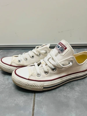 Vita Converse All Star låga sneakers - Klassiska vita Converse All Star sneakers med lågt skaft, vita snören och röd rand längs sulan. Ikonisk logga på plösen och tåhätta i gummi. Tillverkade i canvas för en avslappnad och tidlös look.