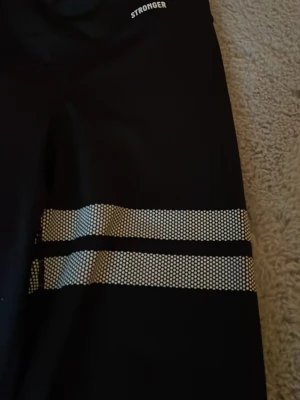 Svarta gympabyxor  från Stronger - Säljer ett par svarta träningsbyxora  från Stronger med vita prickiga ränder runt benen. Tightsen har hög midja och är tillverkade i stretchigt funktionsmaterial som passar perfekt till gymmet eller löpning. Benen är bredare och knte tajta 