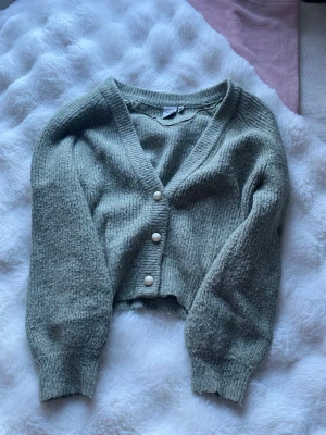 Grön cropped kofta med pärlknappar - Mysig ljusgrön cropped kofta med v-ringning och tre pärlknappar framtill. Koftan har långa ärmar och ribbade muddar vid ärmslut och nederkant. Perfekt för lager på lager och en avslappnad stil.