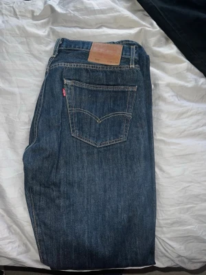 Levi's 501 blå jeans W32 L34 - Klassiska blå Levi's 501 jeans med rak passform och ikonisk röd Levi's-tagg på bakfickan. Jeansen har normal midja och är tillverkade i slitstarkt bomullsdenim. Perfekta för en avslappnad och tidlös stil.