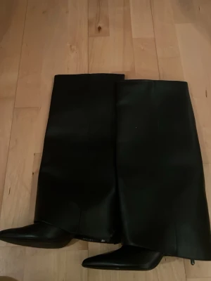 Svarta höga stövlar med spetsig tå - Säljer ett par svarta höga stövlar i skinnimitation med spetsig tå och slät design. Stövlarna har en elegant siluett och passar perfekt till både jeans och kjol. Klassisk look som lyfter vilken outfit som helst. De är lite skadade vid klacken men syns inte mycket på