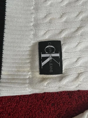 Vit kabelstickad tröja Calvin Klein - Vit kabelstickad tröja från Calvin Klein Jeans med rund halsringning och lång ärm. Tröjan har ett svart CK-märke på nedre delen och är tillverkad i mjukt stickat material. Perfekt för dig som gillar stilrena och klassiska plagg. 