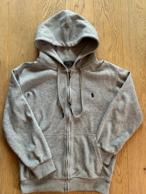 Grå hoodie från Polo Ralph Lauren - Klassisk grå hoodie med dragkedja från Polo Ralph Lauren. , två fickor framtill och den ikoniska broderade loggan på bröstet. Perfekt för en chill och stilren look. Mycket bra skick inga defekter eller hål endast använd fåtal gånger , kan gå ner i pris vid snabb affär. Passar runt 162-172