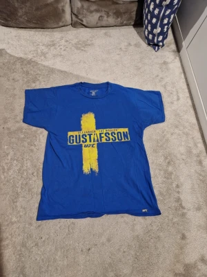 Blå UFC Gustafsson t-shirt - Blå t-shirt från UFC med gult tryck av Alexander Gustafsson och ett kors inspirerat av svenska flaggan. Tillverkad i 100% bomull och har klassisk passform med korta ärmar. Perfekt för dig som gillar MMA och sportig stil.