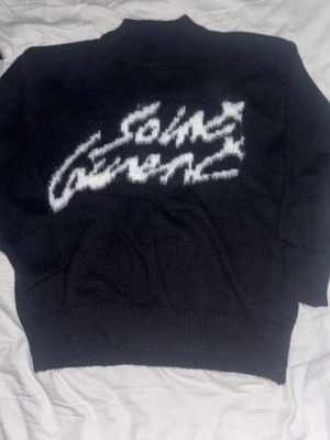 Svart stickad tröja Saint Laurent - Svart stickad tröja med vit Saint Laurent-logga i borstigt mönster över bröstet. Den är ni nyskick i storlek M. Perfekt för dessa kyliga dagar!