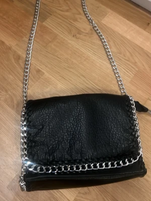 Svart axelväska med kedjedetaljer - Snygg svart axelväska från Tiamo med grov silverfärgad kedja som axelrem och dekor runt locket. Väskan är gjord i skinnimitation med struktur och har ett stort fack samt innerficka med dragkedja. Perfekt för dig som gillar en stockholmstil.