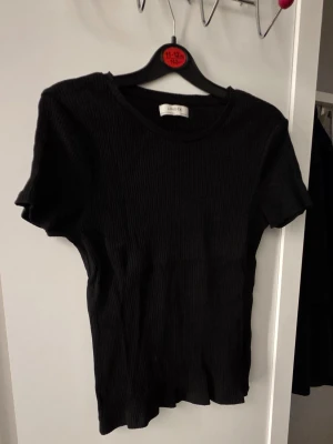 Svart ribbad t-shirt från Lindex - En svart ribbad t-shirt från Lindex med rund halsringning och korta ärmar. T-shirten har en slim passform och är tillverkad i mjuk bomull, perfekt för en stilren och enkel look.