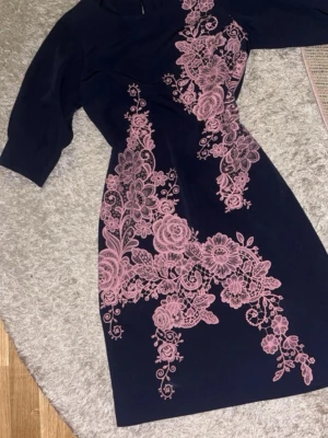Mörkblå klänning med rosa spetsmönster - Supersnygg mörkblå klänning med halvärmade ärmar och detaljerat rosa spetsmönster i blommig design framtill. Klänningen har en figurnära passform och rund halsringning. Perfekt för dig som vill sticka ut med en unik look.